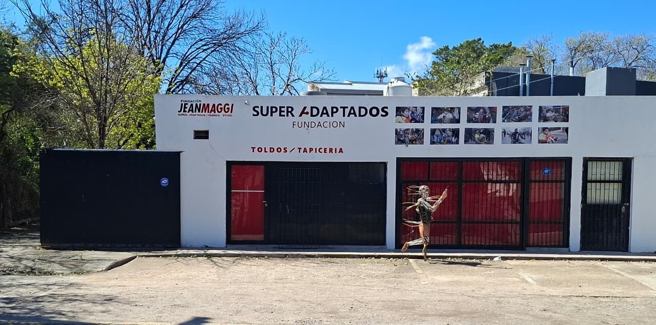 Fachada del edificio de la sede de la fundación Super Adaptados, con paredes blancas, aberturas y portones negros, y el cartel identificatorio con el logotipo en la parte superior